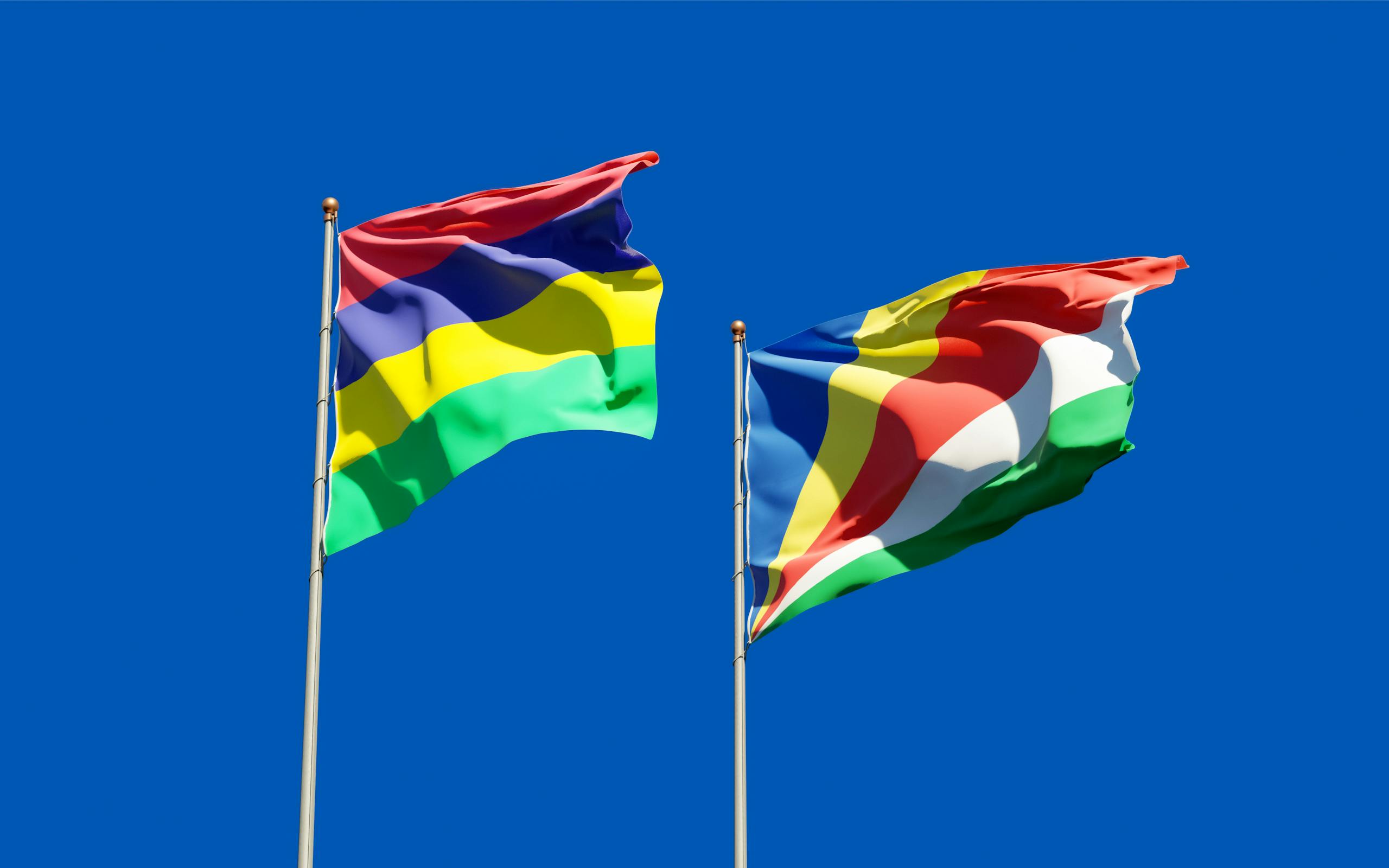 Flags of Mauritius and Seychelles waving on flagpoles with a clear blue sky backdrop, representing unity.